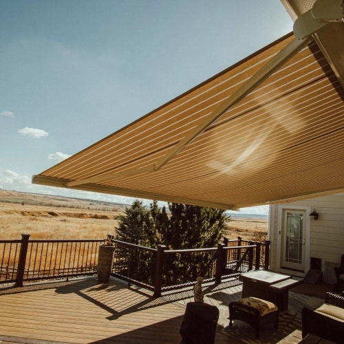A striped retractable awning overhanging a raised wooden porch for a tan home surrounded by a backyard of trees.